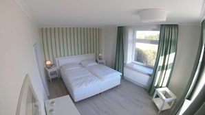 Quarto com cama de casal, mesa de cabeceira com abajur, janela com cortinas e radiador.