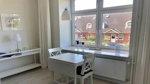 Quarto com mesa e cadeira brancas, um radiador, janela com vista para casas e uma estante com luminária.