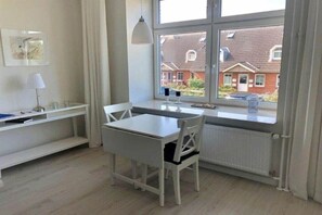 Ein Zimmer mit einem weißen Tisch und Stuhl, einem Heizkörper, einem Fenster mit Blick auf Häuser und einem Regal mit einer Lampe.