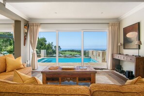 Amplia sala de estar con un gran ventanal corredera que da a una zona de piscina, una mesa de centro de madera y un sofá con cojines.