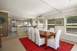 A modern kitchen with white cabinetry, a central island, and a dining area with a wooden table and white chairs. - Stunning house on Pezula Championship Course, 4 bedrooms ensuite, private pool (Knysna)