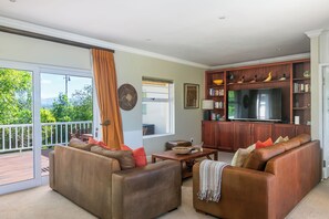 A spacious living room with a large flat-screen TV, leather sofas, a wooden bookshelf, and a balcony with a view of greenery. - Stunning house on Pezula Championship Course, 4 bedrooms ensuite, private pool (Knysna)