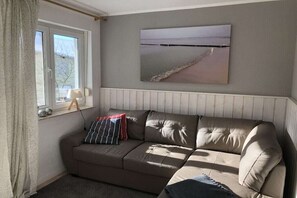Ein Wohnzimmer mit einem beigen Sofa, einem Vorhangfenster, einer Lampe und einem Gemälde mit einer Strandansicht.