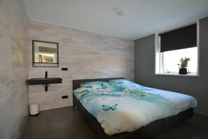 Een slaapkamer met een bed, een nachtkastje, een wastafel aan de muur en een raam met uitzicht op het omliggende landschap.