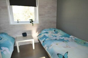 Ein Schlafzimmer mit zwei Einzelbetten, einem kleinen weißen Tisch, einem Fenster mit einer Pflanze und Blick ins Freie.
