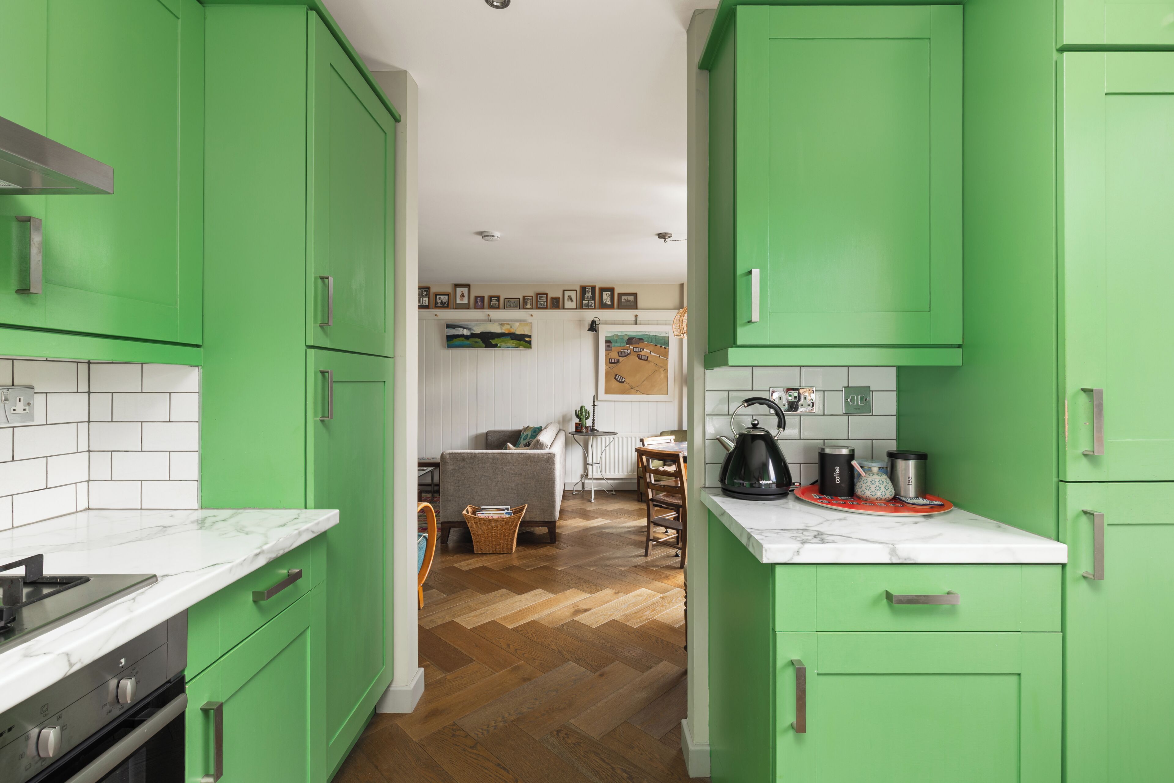 Une cuisine avec des meubles de couleur verte, un plan de travail en marbre et un parquet en bois disposé en chevrons.