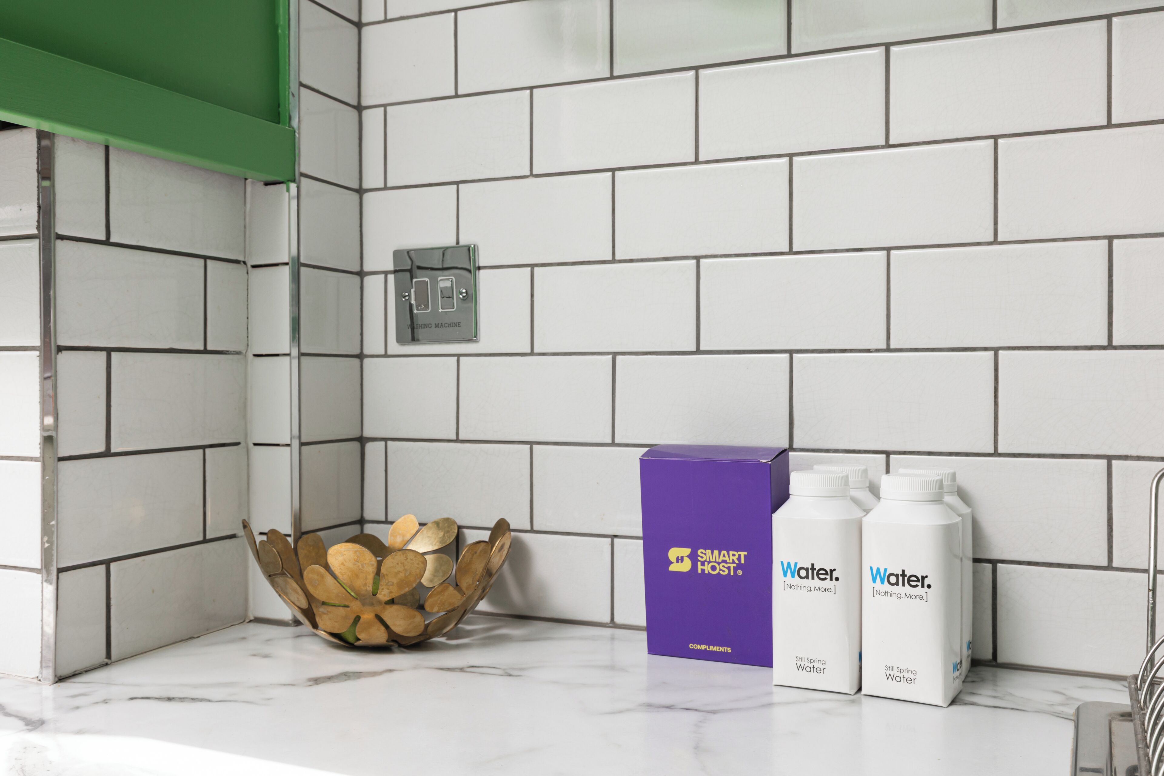 A bathroom with white tiled walls, a green shower curtain, a washing machine, a wooden bowl, and two bottles of water.