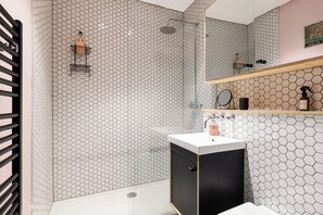A modern bathroom with a hexagonal tile pattern, a shower with a glass door, a black cabinet with a white countertop, and a wooden shelf with toiletries. - New! Delightful 2BR Near Imperial Wharf & Chelsea (Greater London)