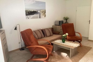 Ein Wohnzimmer mit einer Couch, zwei Sesseln, einem Couchtisch mit einer Vase, einem Bild an der Wand und einer Tür.
