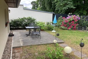 Eine Terrasse mit Tisch und Stühlen, umgeben von Pflanzen und einem blauen Sonnenschirm.