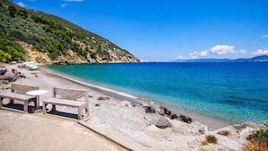 A beach with clear turquoise water, a rocky shoreline, and a hilly backdrop.