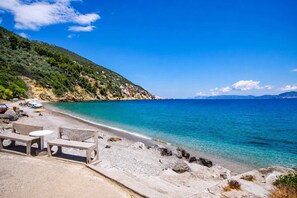 A beach with clear turquoise water, a rocky shoreline, and a hilly backdrop.