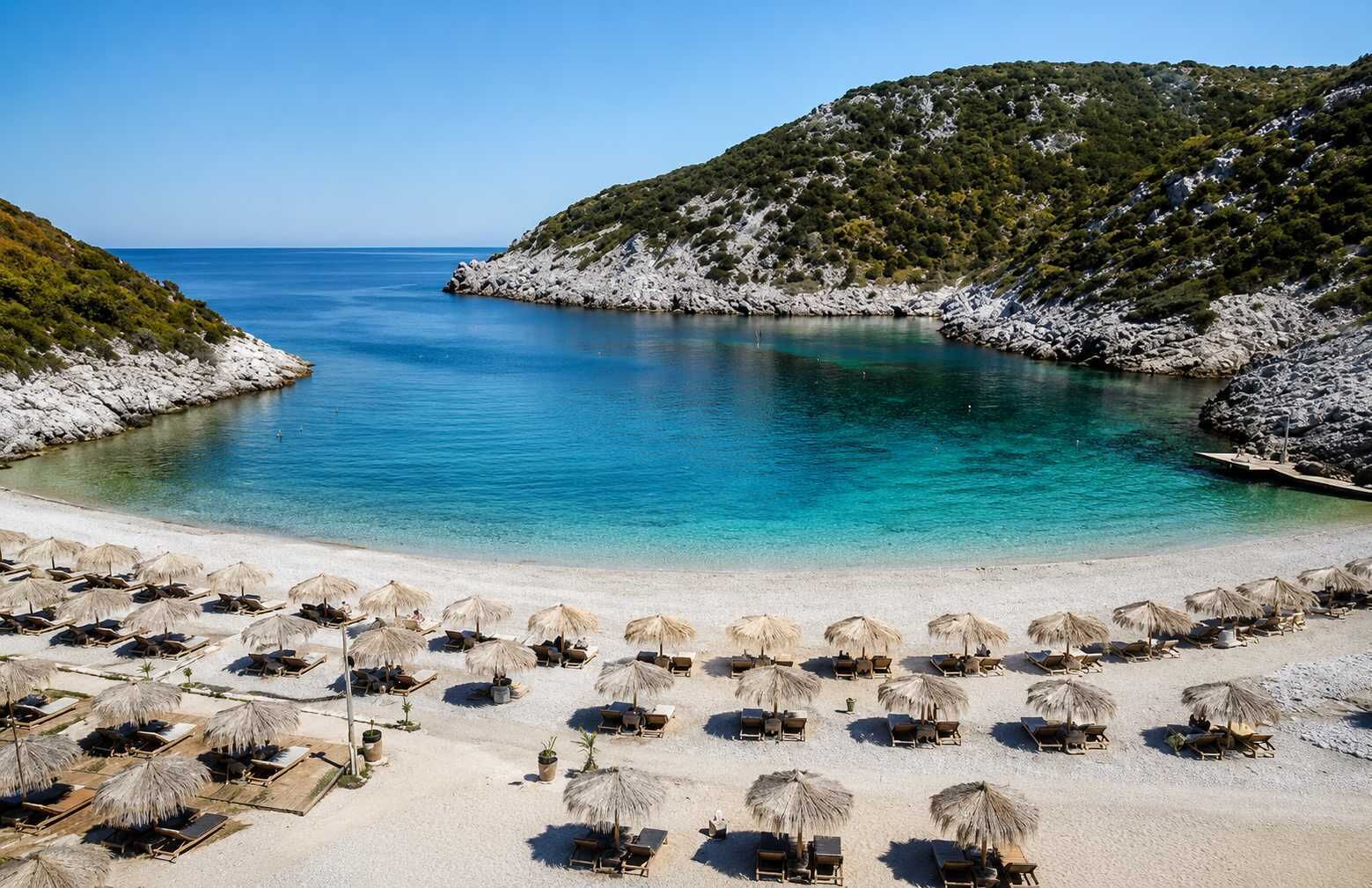 Una playa con agua turquesa cristalina, arena blanca y una colina rocosa al fondo. Se ven filas de tumbonas y sombrillas de paja.