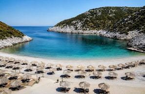 A beach with clear turquoise water, white sand, and a rocky hill in the background. There are rows of sun loungers and thatched umbrellas.