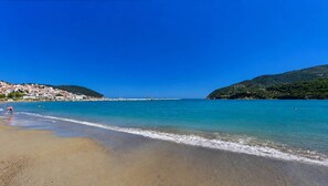 A beach with clear blue water, a sandy shore, and a backdrop of hills with buildings.