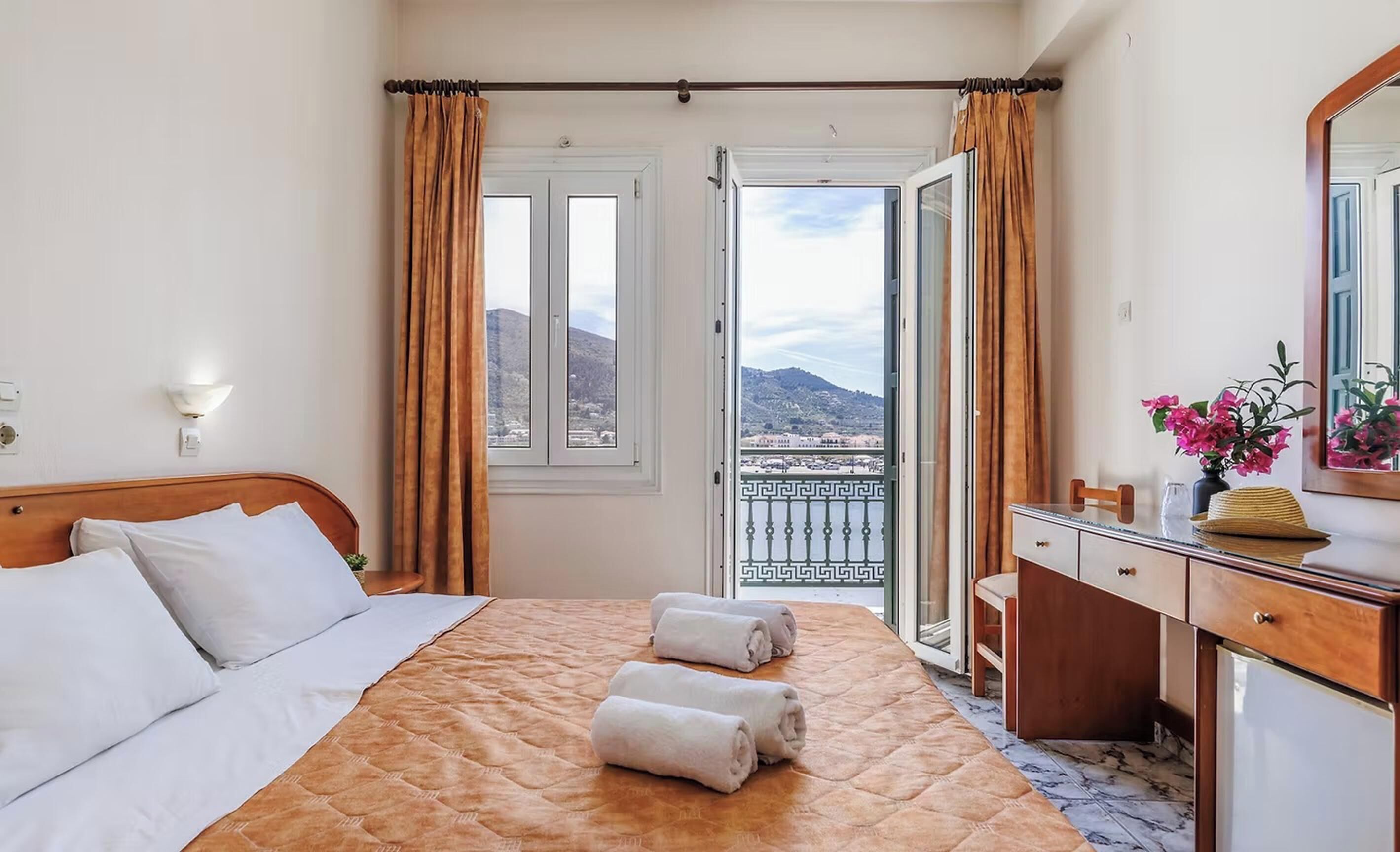 A hotel room with a bed, a desk, a chair, a mirror, and a view of the city and mountains through the window.