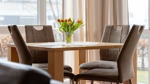 Uma sala de jantar com mesa posta para dois, cadeiras, um vaso com flores e vista pela janela.