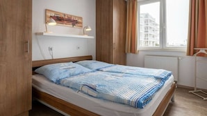 Quarto com cama, guarda-roupa de madeira, uma estante com abajur e janela com cortinas.