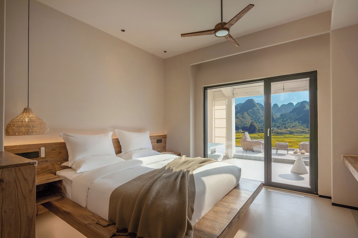 Un dormitorio con una cama grande, un ventilador de techo, vistas a montañas a través de una puerta corrediza y una mesita de noche de madera.
