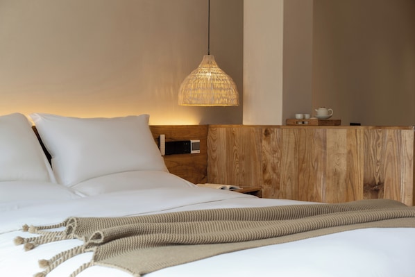 A neatly made bed with white linens, a wooden headboard, a bedside table with a lamp, and a small tray with cups and a teapot.