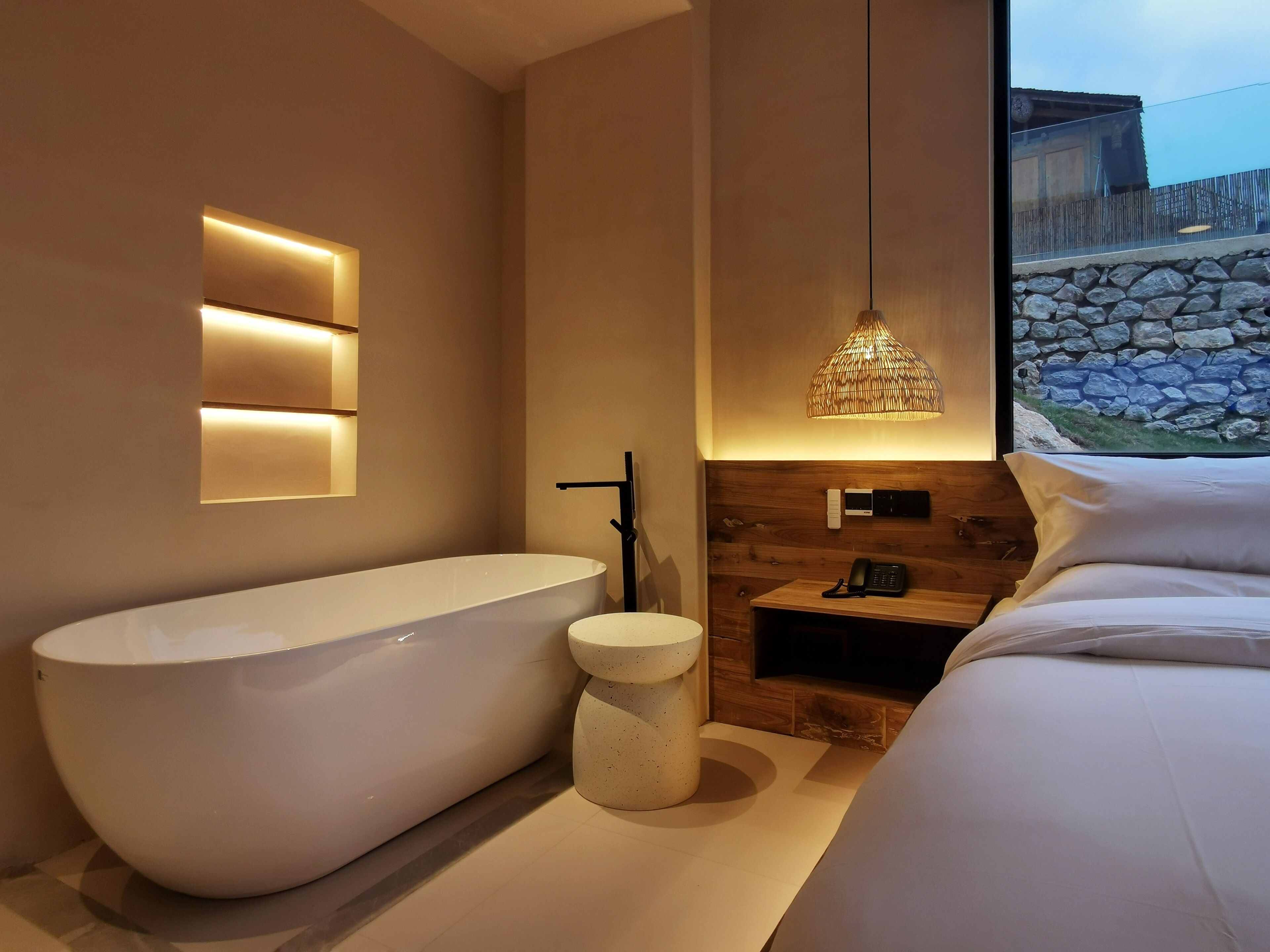 A modern bathroom with a freestanding bathtub, a small round sink, and a bedside table with a phone.