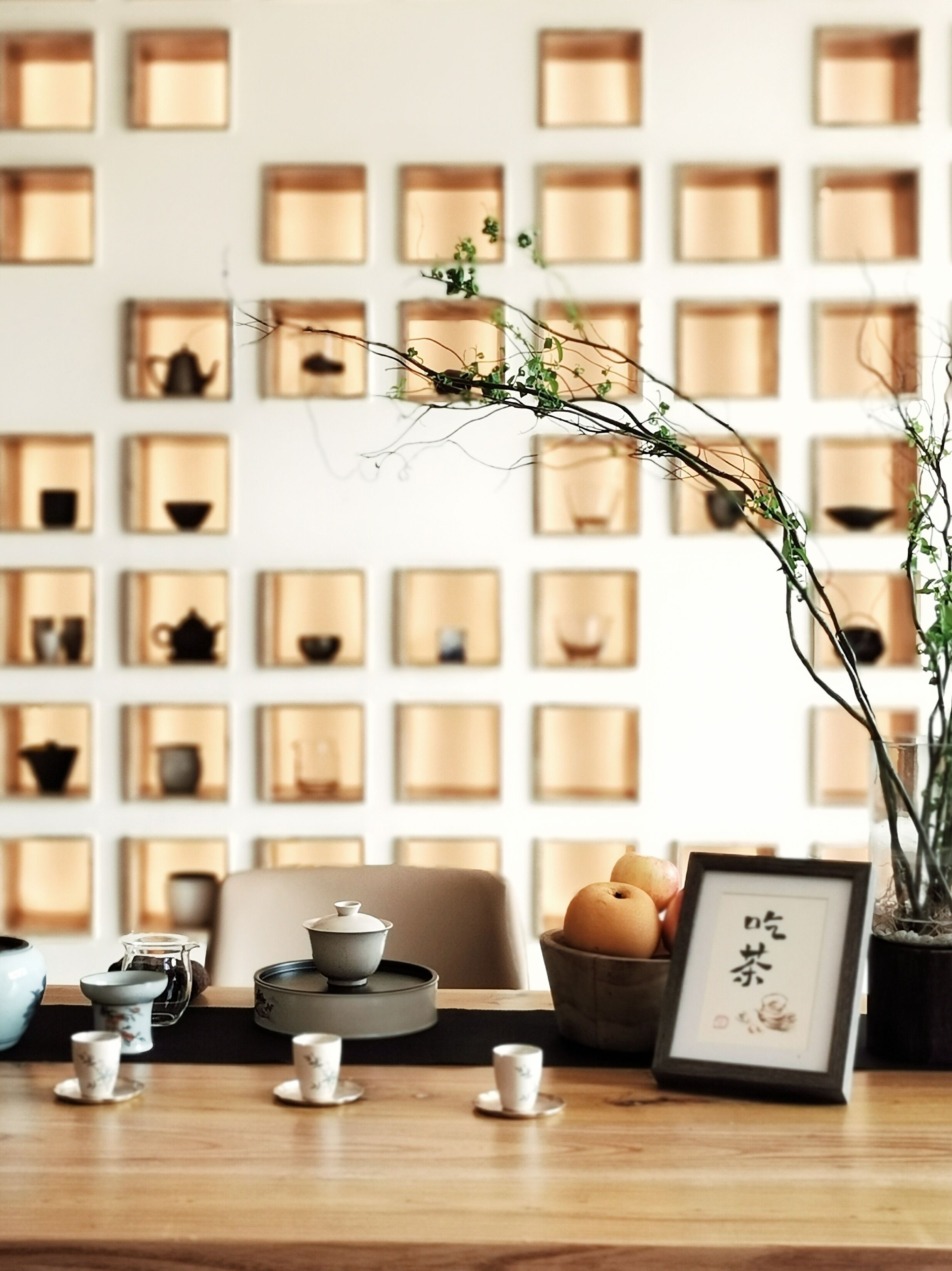 Una habitación con una mesa de madera, un juego de té, un cuadro enmarcado y una pared con estanterías que contienen varios objetos.