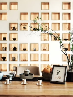 A room with a wooden table, a tea set, a framed picture, and a wall with shelves containing various items.