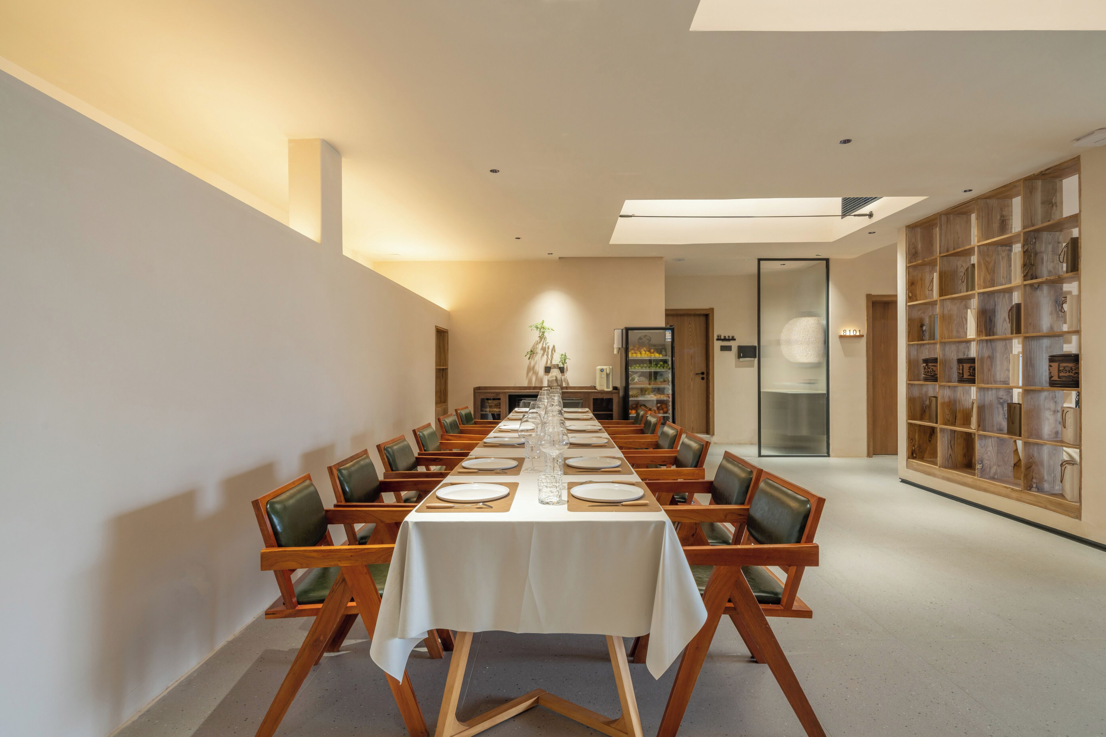 Una larga mesa de comedor con sillas, mantel blanco y una estantería con repisas de madera.