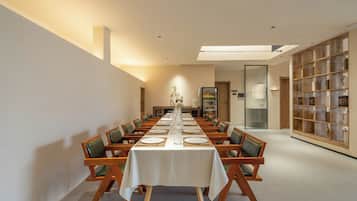 Una larga mesa de comedor con sillas, mantel blanco y una estantería con repisas de madera.