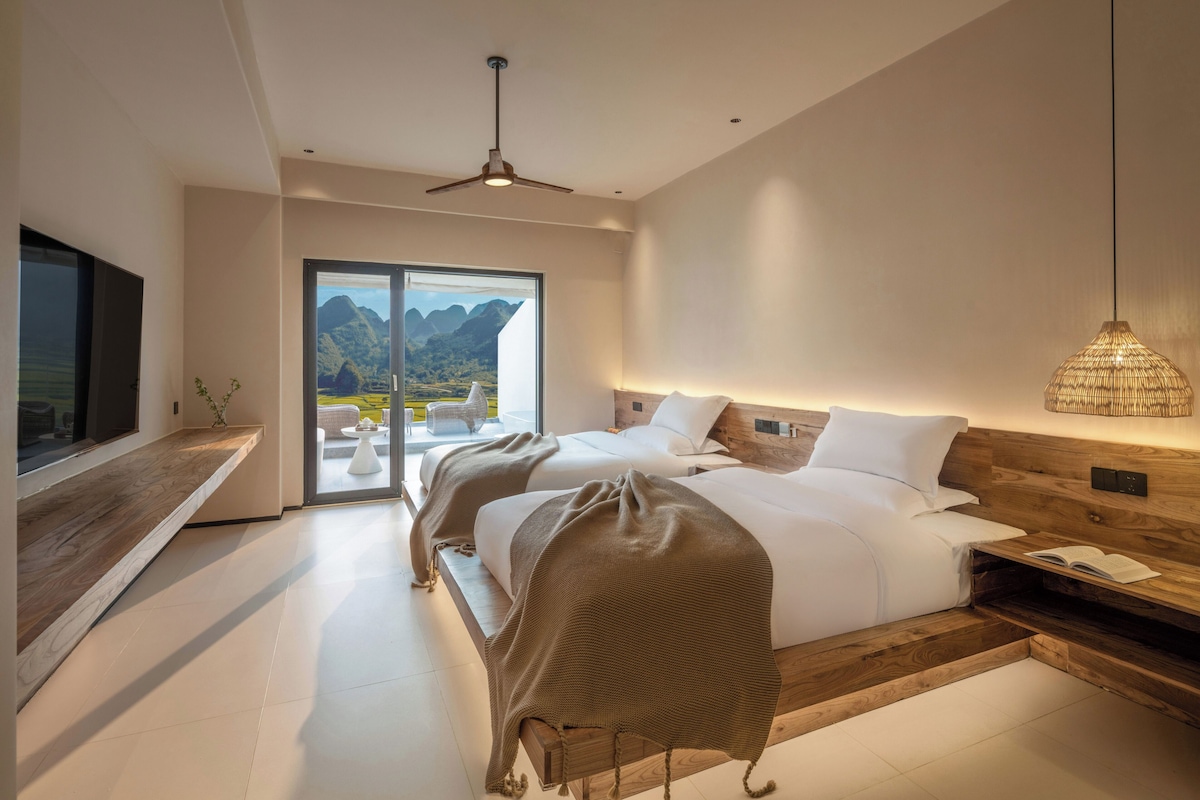 Un dormitorio moderno con una cama grande, una mesita de noche de madera, un televisor y vistas a montañas a través de un ventanal.