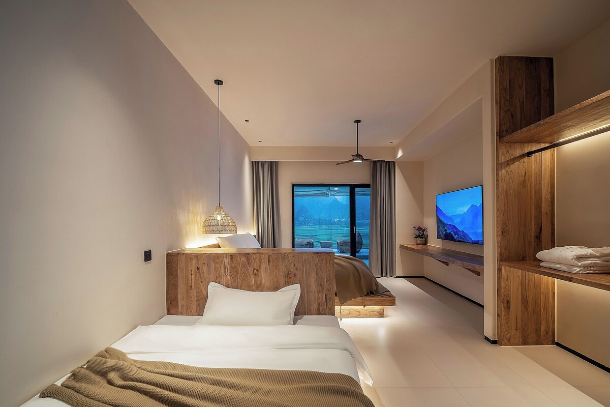 Habitación de hotel moderna con cama, televisor, cabecero de madera y vistas al exterior.
