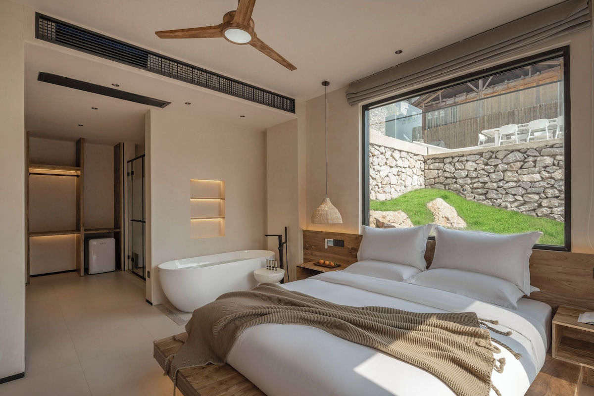 Un dormitorio moderno con una cama grande, una bañera y vistas a una pared de piedra y vegetación.