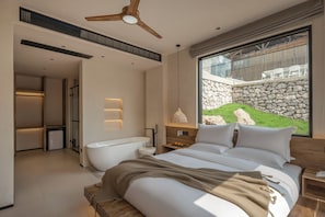 Un dormitorio moderno con una cama grande, una bañera y vistas a una pared de piedra y vegetación.