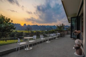 Una terraza con sillas y mesas blancas, con vistas a un paisaje montañoso al atardecer.