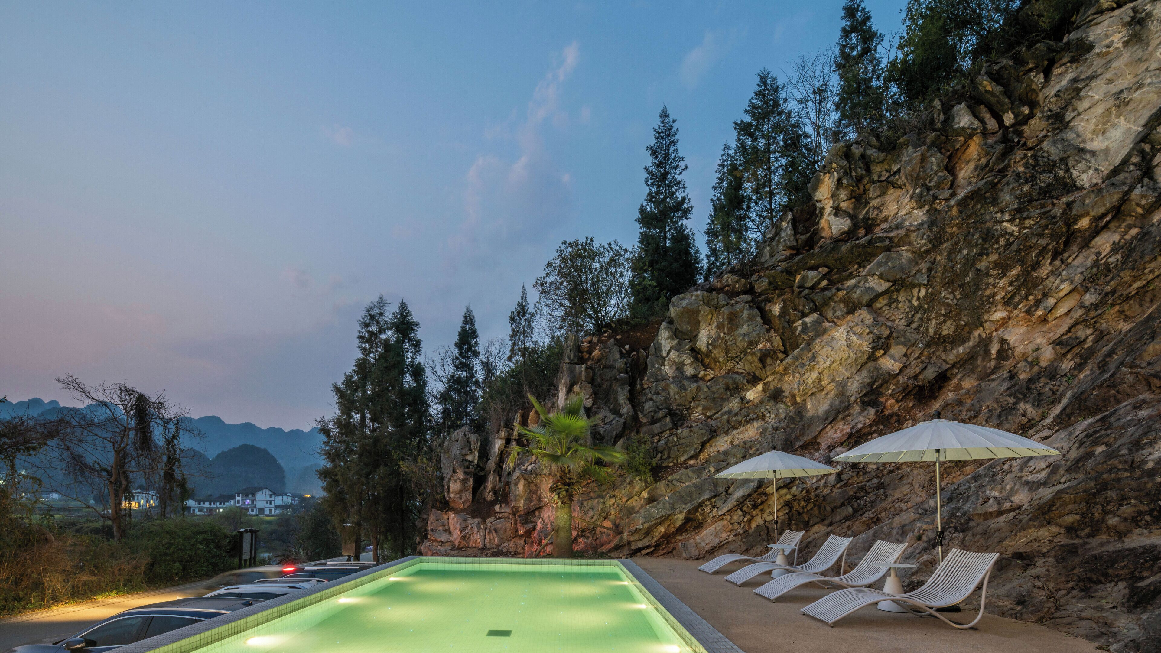 A swimming pool with lounge chairs and umbrellas, surrounded by rocky terrain and trees.