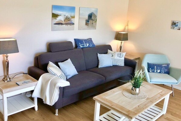 Een moderne woonkamer met een grijze bank, een houten salontafel en een lichtblauwe fauteuil. Er staan twee bijzettafels met lampen bij, en de muren zijn versierd met ingelijste foto’s.