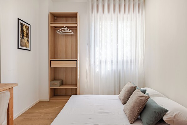 A bedroom with a bed, pillows, a wooden wardrobe, and a window with sheer curtains.
