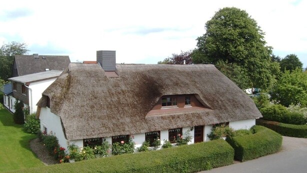 Uma casa de campo tradicional com telhado de palha e fachada branca, cercada por sebes bem cuidadas e um gramado.