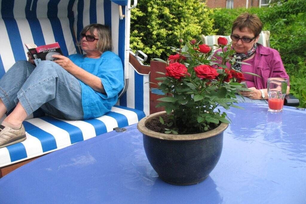 Dos personas sentadas en un banco azul y blanco a rayas, una leyendo un libro y la otra con una bebida. Una planta grande en maceta con flores rojas está en primer plano.