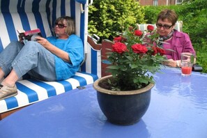 Zwei Personen sitzen auf einer blau-weiß gestreiften Bank, eine liest ein Buch und die andere hält ein Getränk. Im Vordergrund steht ein großer Topf mit roten Blüten.