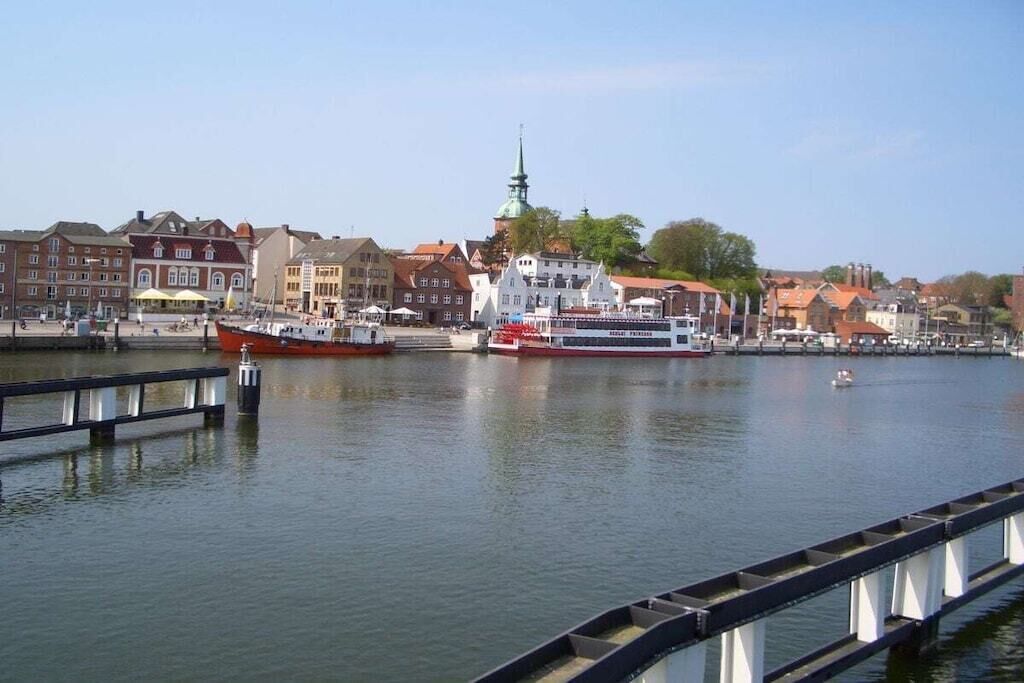 Une promenade en bord de mer avec un bateau rouge amarré, un traversier blanc et rouge, et des bâtiments à l’architecture traditionnelle.