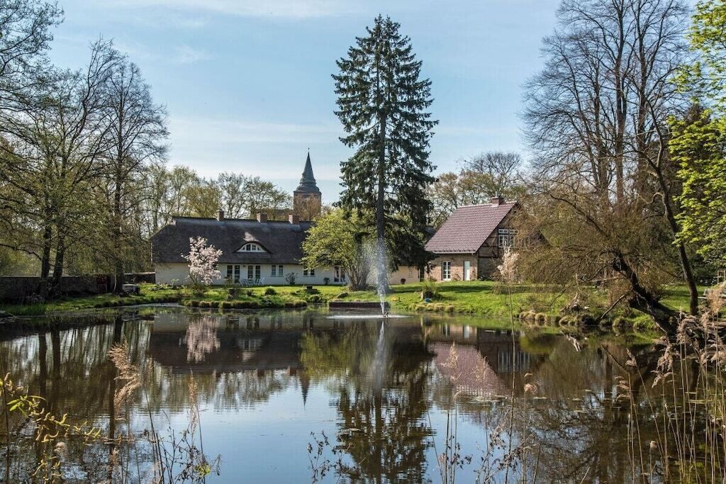 Un étang paisible avec une fontaine, entouré d’arbres et de bâtiments, dont un clocher d’église particulièrement imposant.