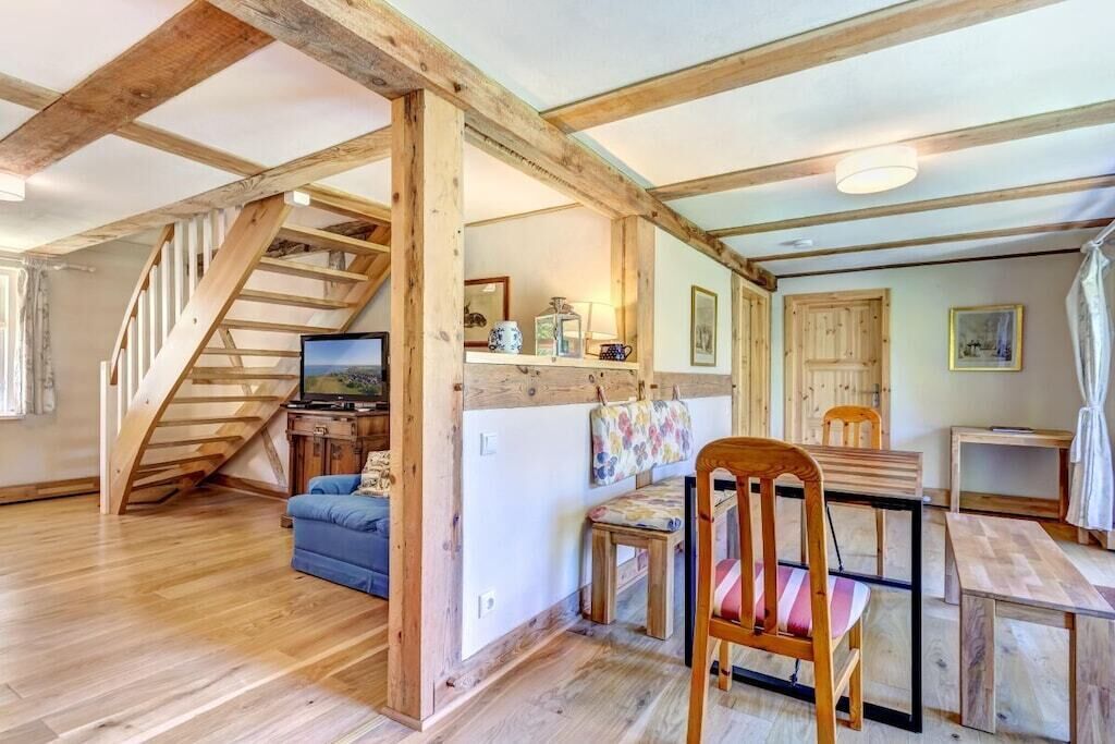 Een kamer met een houten trap, een televisie, een eettafel met stoelen en een klein keukentje.
