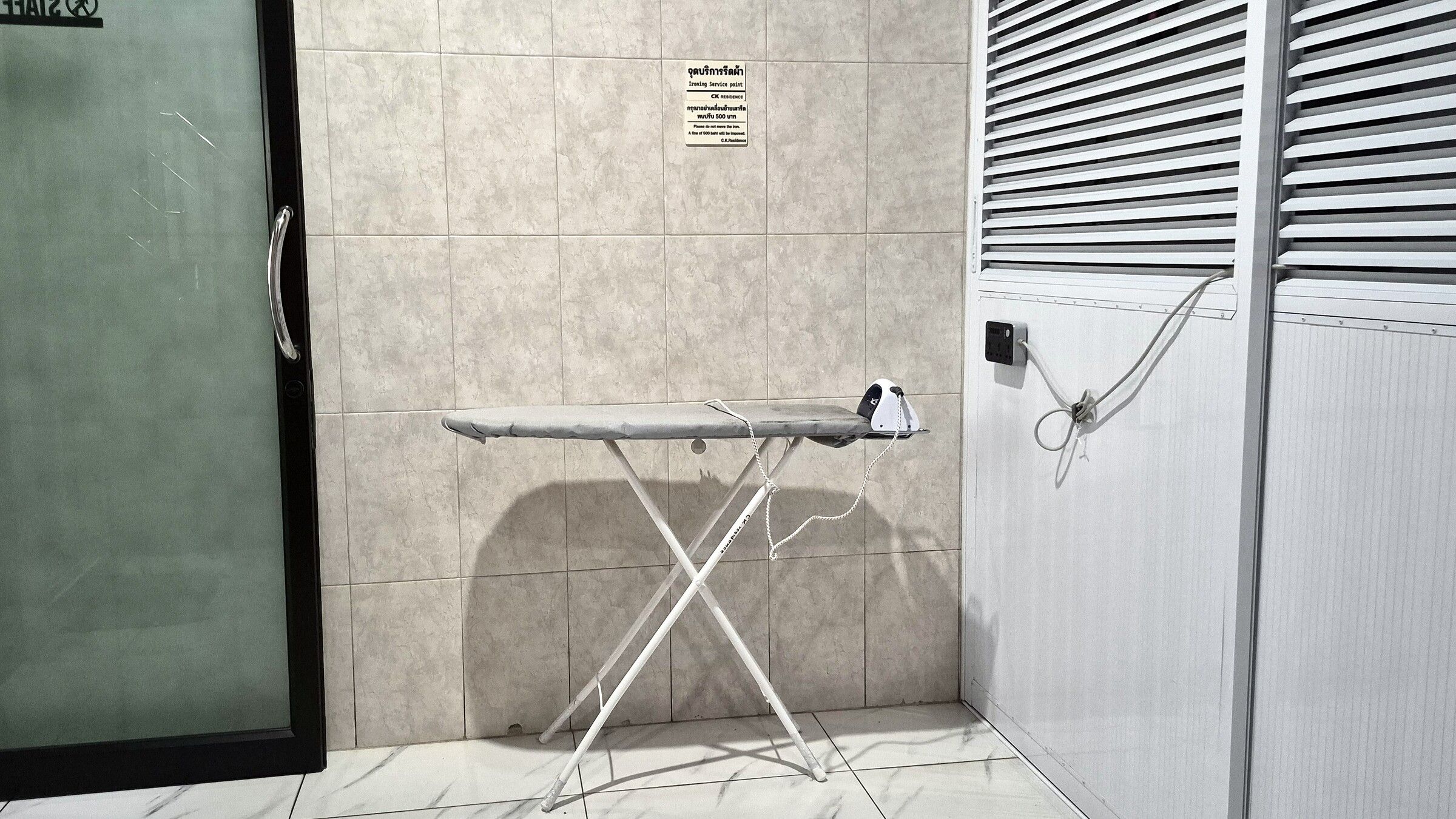 Una habitación con piso de baldosas, una tabla de planchar metálica y un enchufe eléctrico en la pared.