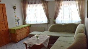 A living room with a sofa, a wooden cabinet, a small table with a glass, and a window with curtains. - Holiday apartment with 1 bedroom (Tönning)