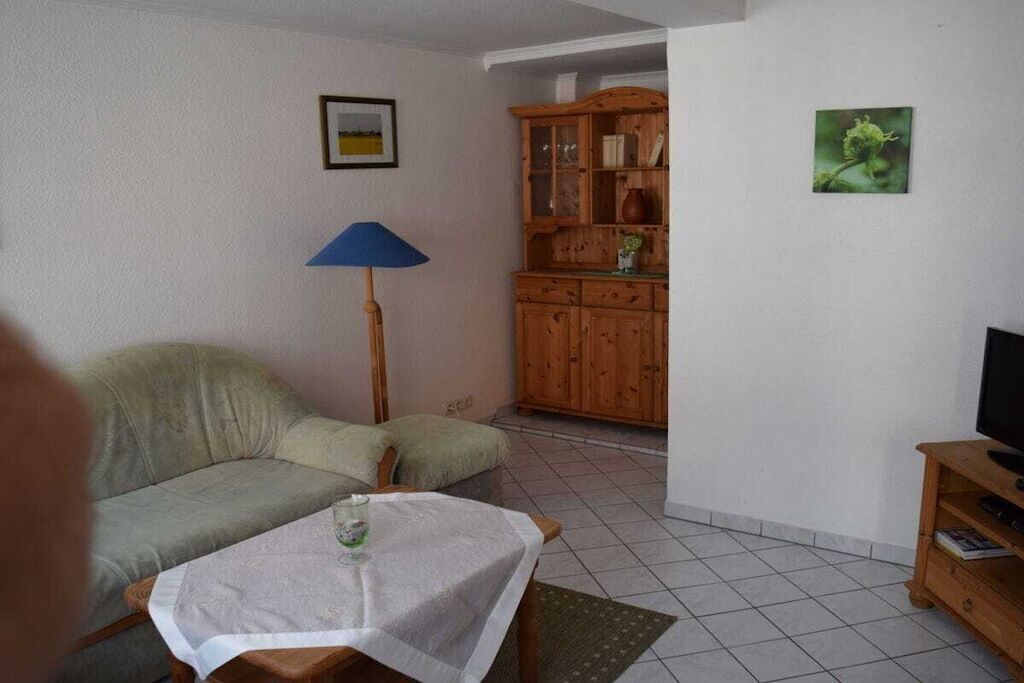 Uma sala de estar com sofá, mesa com um copo, luminária, armário de madeira e televisão sobre um suporte.