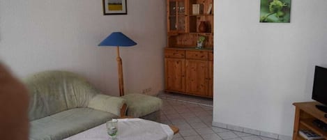 Uma sala de estar com sofá, mesa com um copo, luminária, armário de madeira e televisão sobre um suporte.