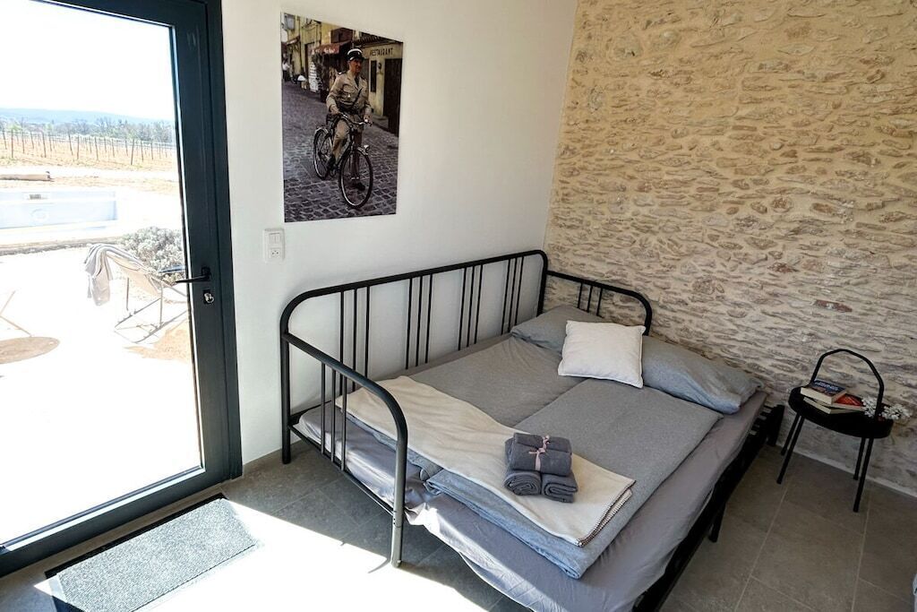 Een enkel bed met metalen frame, een klein nachtkastje, een stenen muur en uitzicht op een straat.