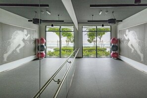 A modern gym with mirrored walls, exercise equipment, and large windows offering a view of greenery and a body of water. - Luxe 1 Bed l Private Beach Access & City Views (Emaar Beachfront)