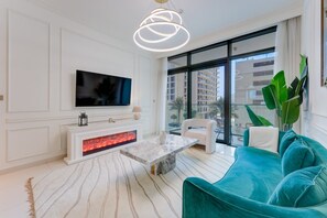 A modern living room with a flat-screen TV, a fireplace, a sofa, and a large window offering a view of buildings. - Luxe 1 Bed l Private Beach Access & City Views (Emaar Beachfront)